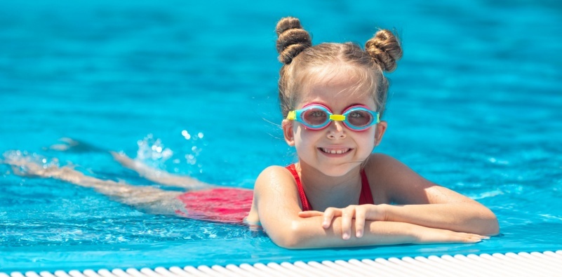 A young girl wearing swimming goggles, holding on to the side of an outdoor swimming pool.